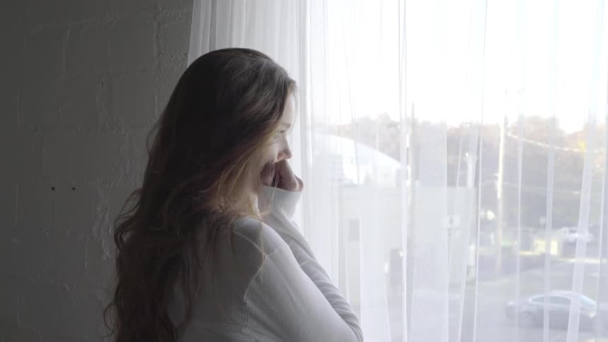 Woman rests her hand on her fist as she looks out the curtained window