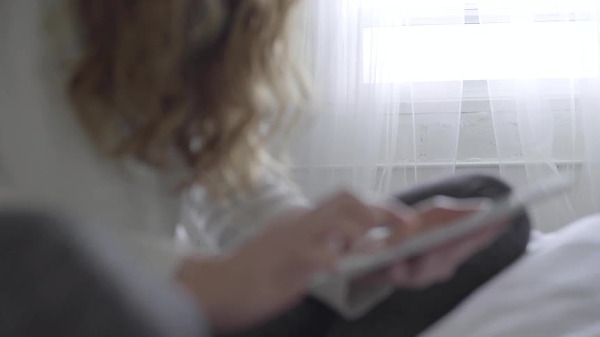 A woman scrolls on a tablet while sitting on a bed