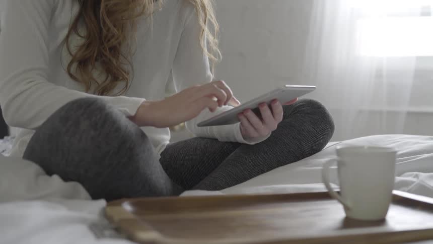 A white woman scrolls through her smartphone while sitting on a bed