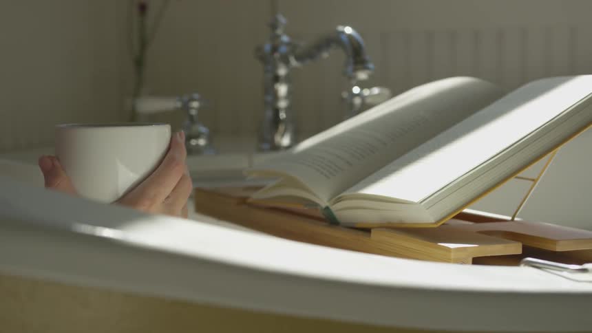 A person reads a book and drinks from a mug while taking a bath