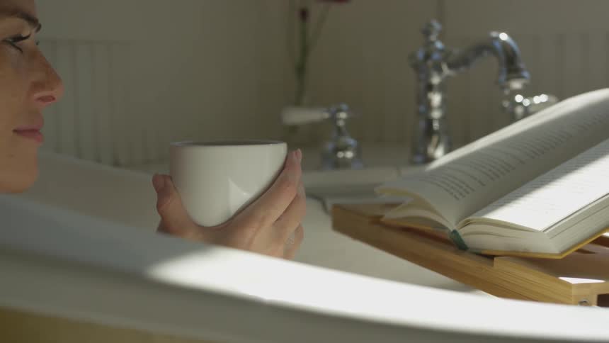 A white female reads a book and holds a coffee mug while soaking in a bathtub