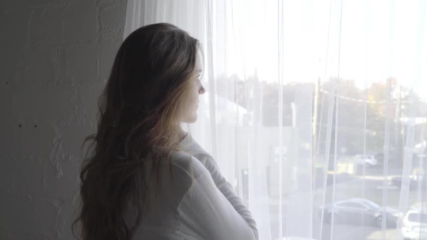A woman stands by a window and looks out of it