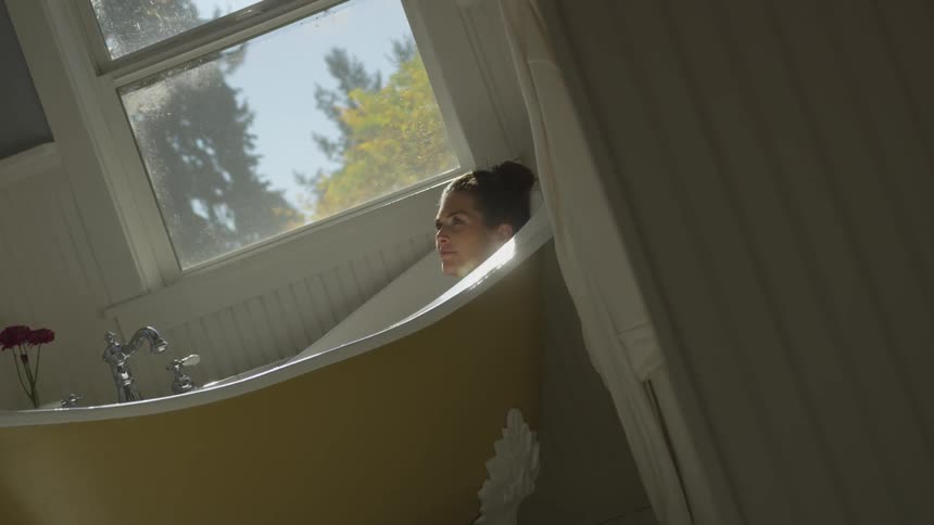 A woman relaxes in a deep bathtub next to a window