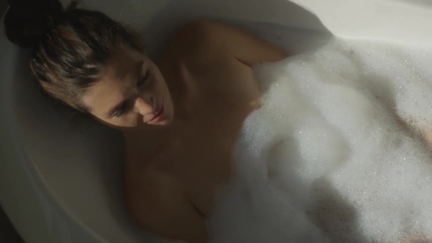 A woman sits and relaxes in a bathtub filled with bubbles covering her unclothed body