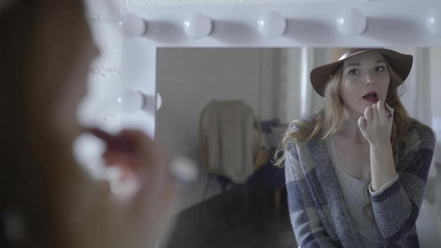 A young woman wearing a faux cowboy hat sits in front of a vanity mirror while she thoroughly applies deep red lipstick to her lips