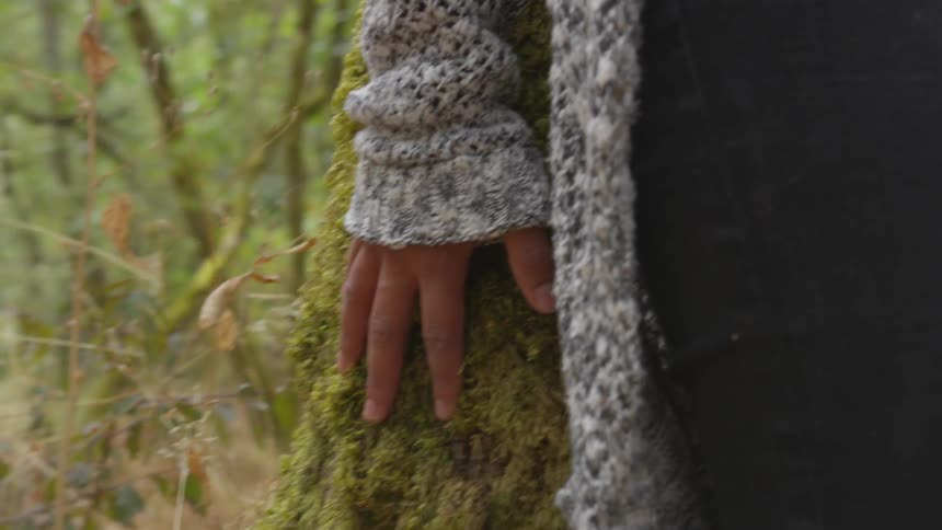 A woman's hand touches a moss covered tree trunk as she hides behind it