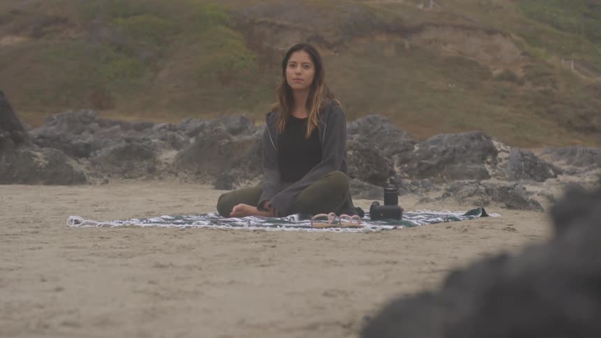 A young woman sitting on a towel at a beach with a camera and sandals