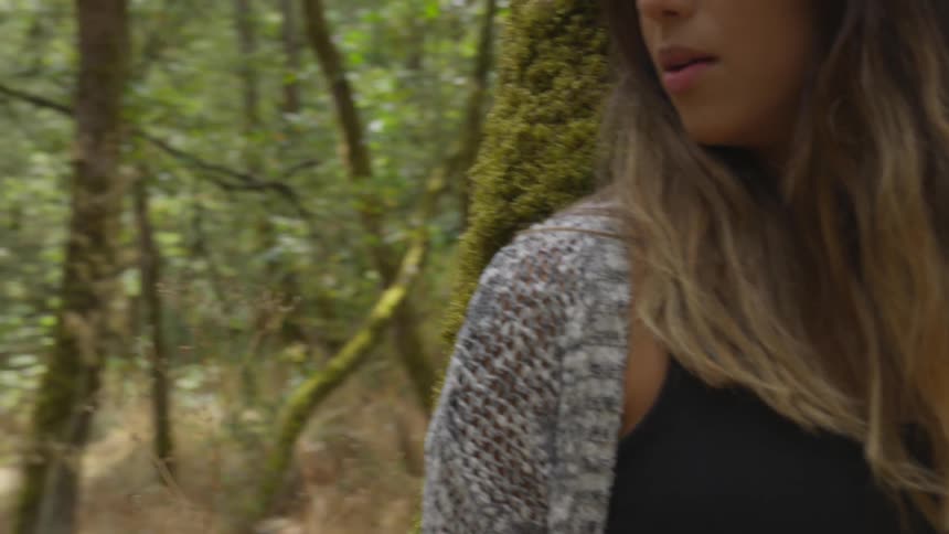A white woman breathes heavily while hiding behind a moss covered tree