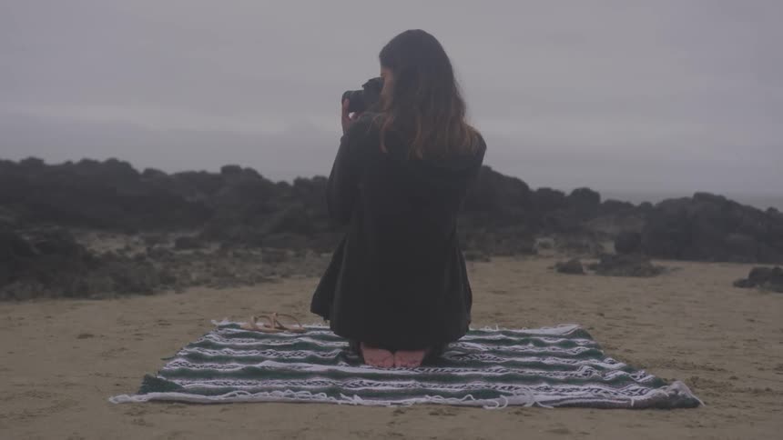The woman is sitting on a blanket at the beach taking pictures of the rocks on an overcast day