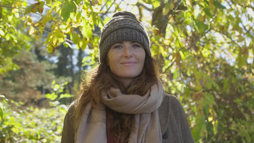 An older woman in a knit cap and wearing a scarf smiles in a forest