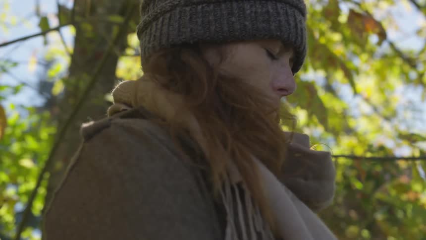 A woman in a jacket and scarf stands beneath a tree with her eyes closed