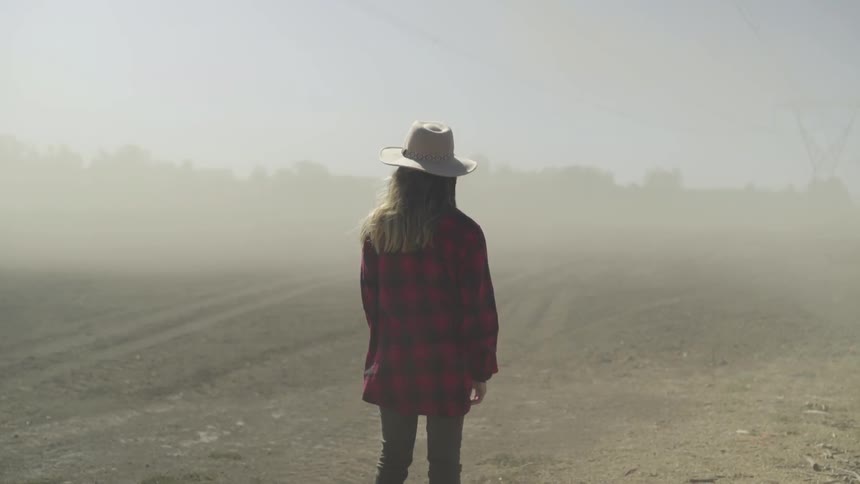 Young woman in cowboy hat faces away from camera in dusty field