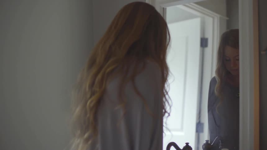 A white woman stands in front of a mirror and looks down