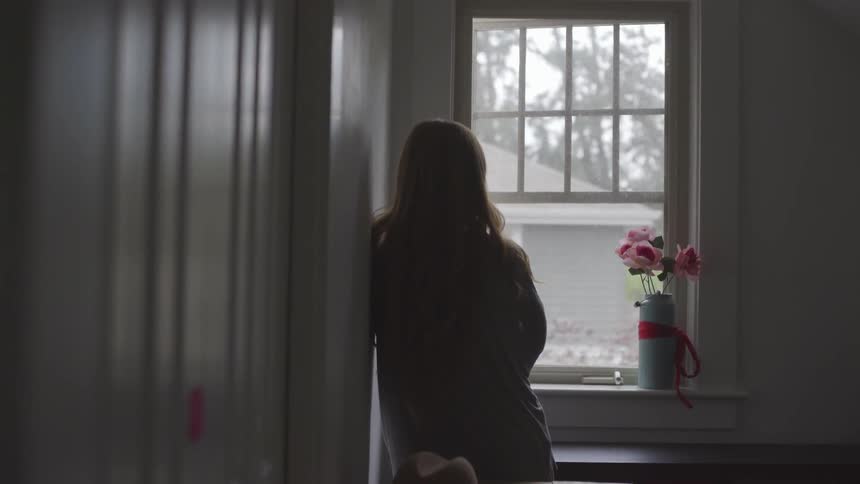 A female leans against a wall while looking out a window with a vase of flowers in front of it