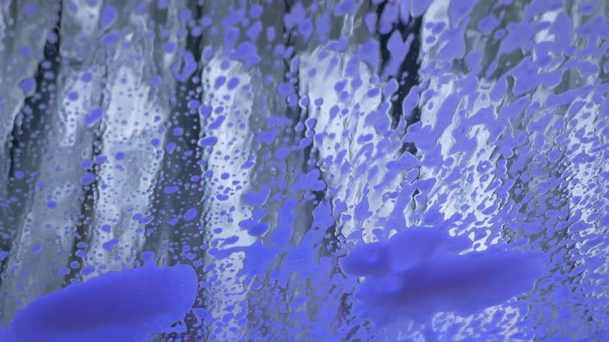 A windshield is washed as a light appears to change the colors of the soap bubbles