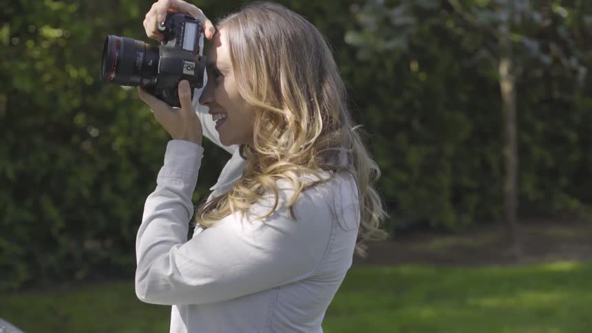 A white female smiles as she takes pictures with a camera in a nature setting