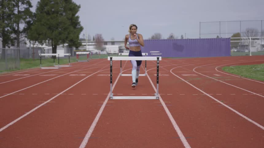 A woman wearing athletic gear runs and jumps over a hurdle on a racing track