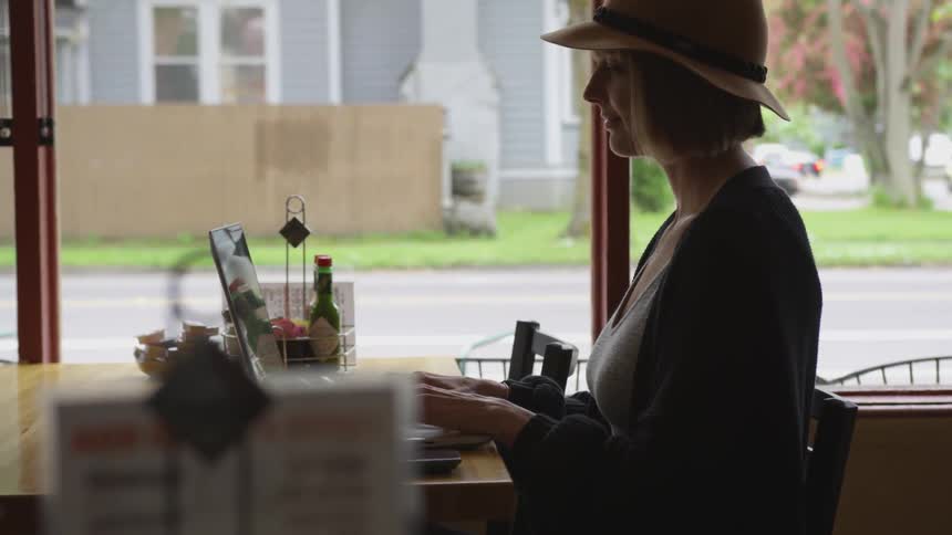 A woman sits at a restaurant table near large windows working on her laptop