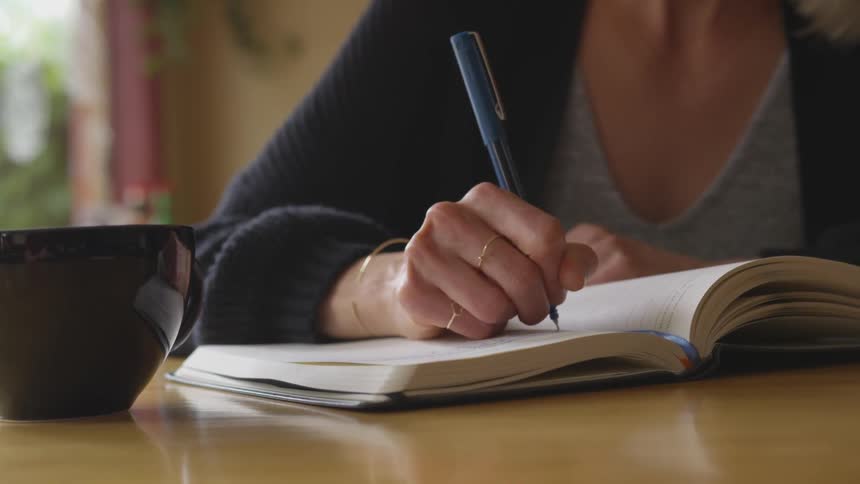 A woman writes in a notebook while sitting at a table with a cup of coffee
