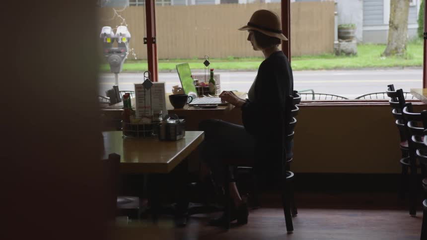 A white woman wearing a hat sits at a table in a restaurant typing on her laptop