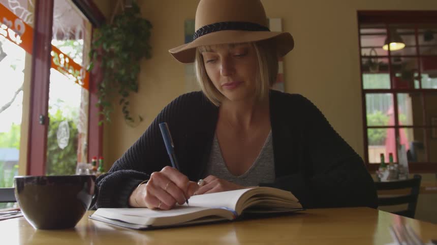 A woman in a hat writes in a notebook while sitting in a coffee shop