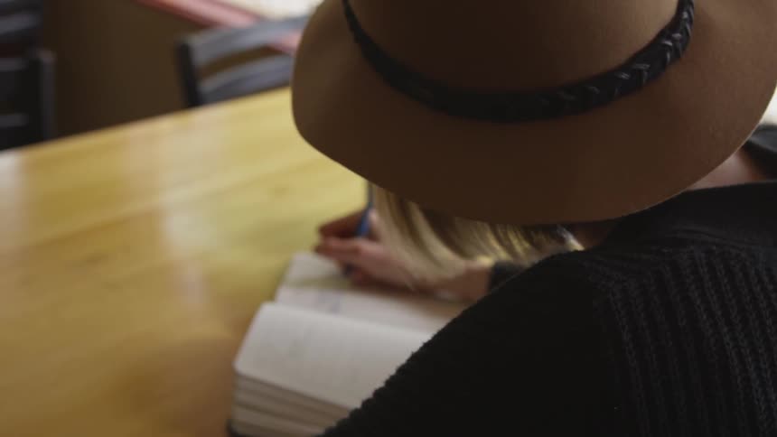 Woman in hat sits at desk and writes in journal with pen
