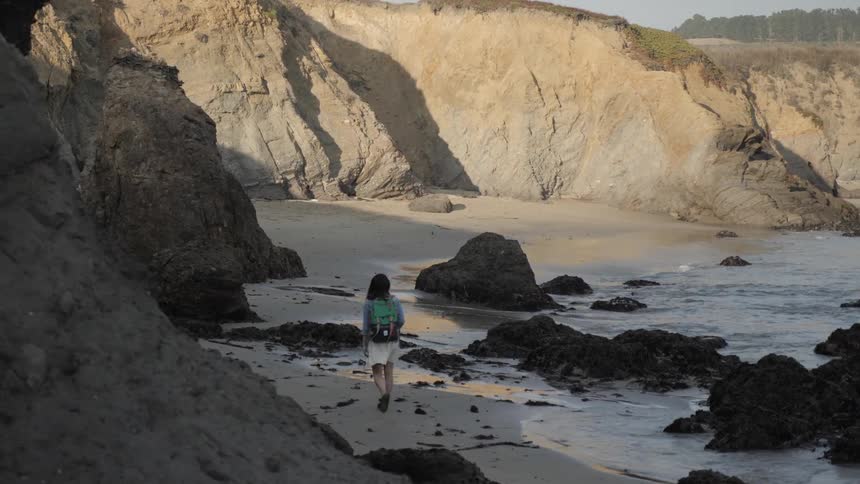 A woman with a backpack walks on a beach alongside a cliff