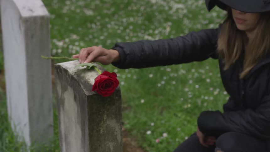 A woman dressed in black places a red rose on a headstone in a cemetery