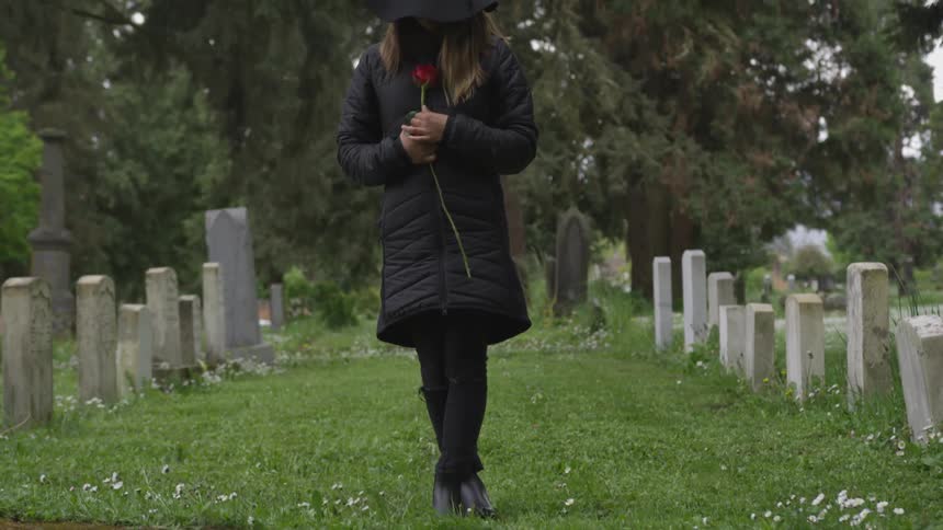 A woman wearing black and holding a red rose stands with her head bowed in a cemetery
