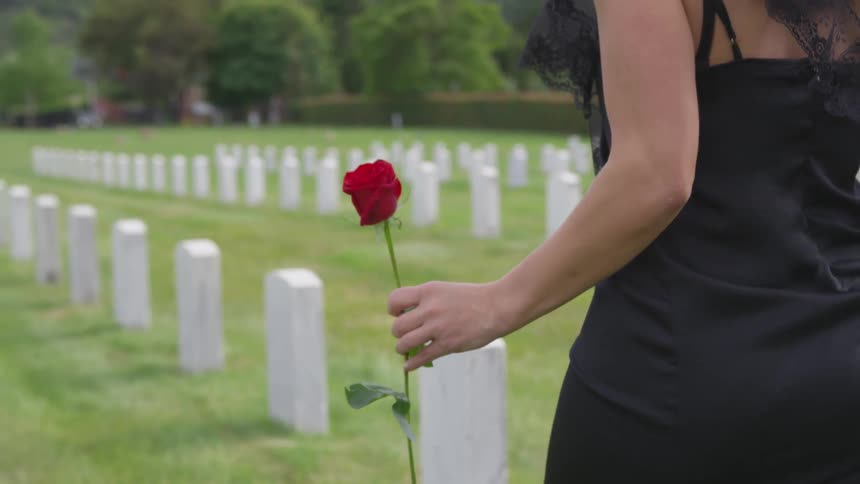 A young widow dressed in black and carrying a rose walks between the rows of white headstones in a cemetery
