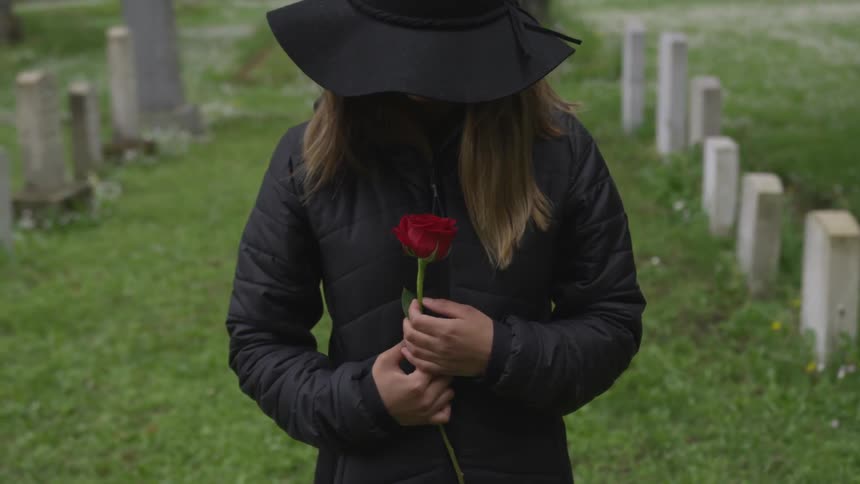 A woman holds a rose to her chest at a graveyard