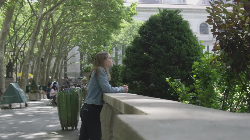A young white woman leans against a wall and looks at a tree