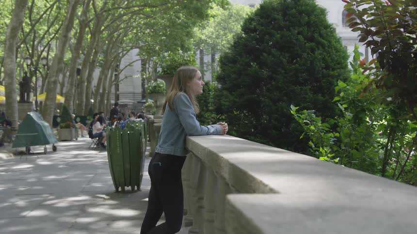 A white female leans against a low cement wall at a park with trees and shrubs around her