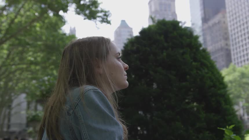 Blonde woman sits down in public park and looks at the tall buildings in the daytime
