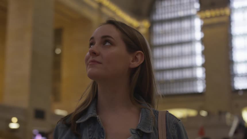 A young woman stands in grand central terminal in new york city and she is looking around toward the ceiling taking in the impressive architecture