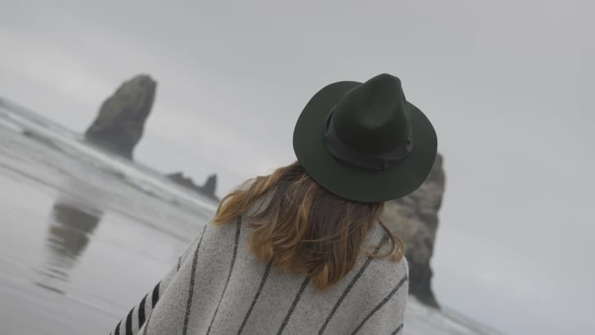A woman in a black hat and grey poncho stands on a beach and stares at the water