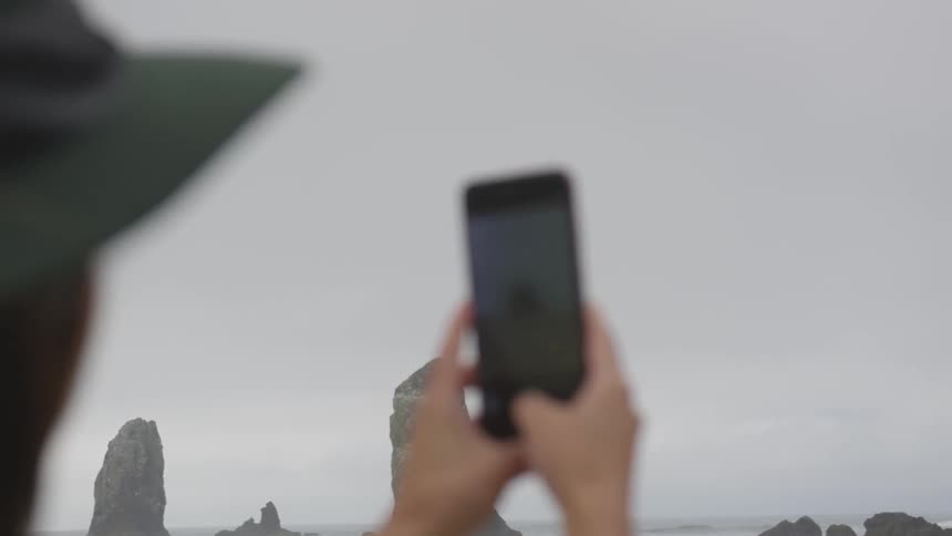 A woman wearing her hat takes a photo at the beach with her smartphone