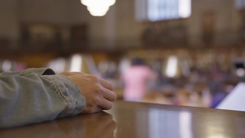 The arms are laying across the wooden table with the hands close with denim sleeves the background is fuzzy but it looks like some kind of classroom and a teacher maybe an exercise class or music class
