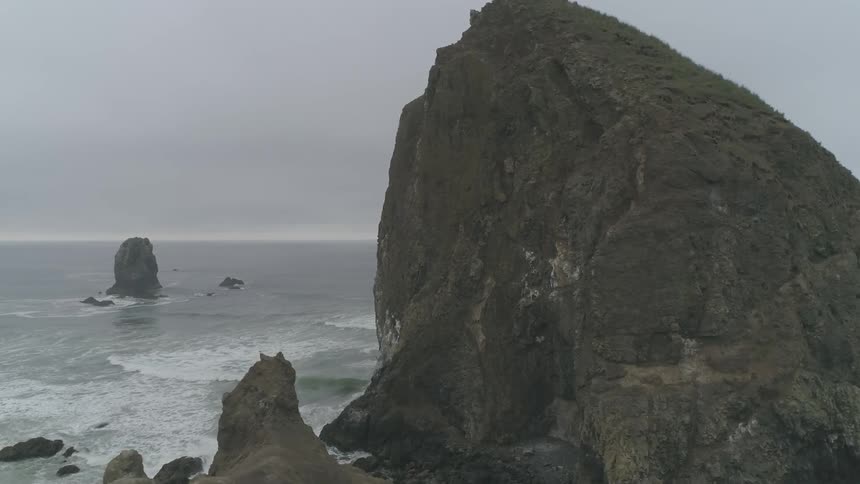 The camera looks at a mass of rock coming out of the ocean