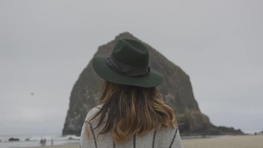 Woman standing on a beach looking out toward the water and a large rock