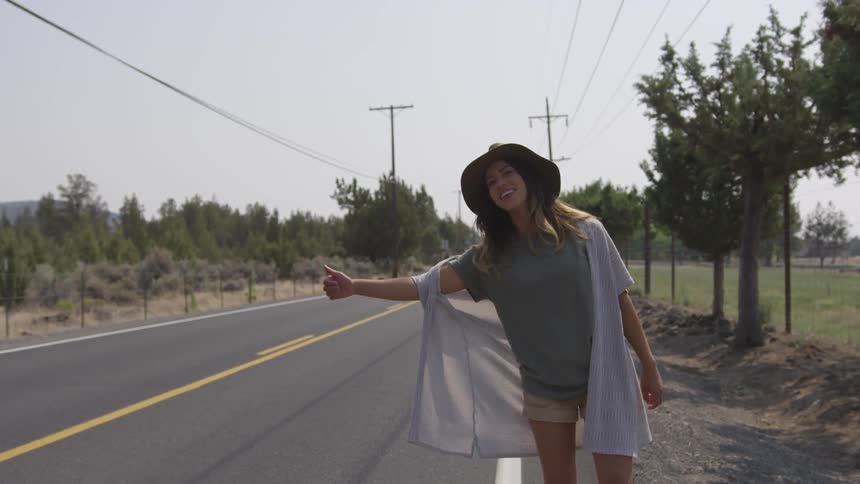A young white woman wearing a hat and a thin overcoat attempts to hitch a ride from the side of a road