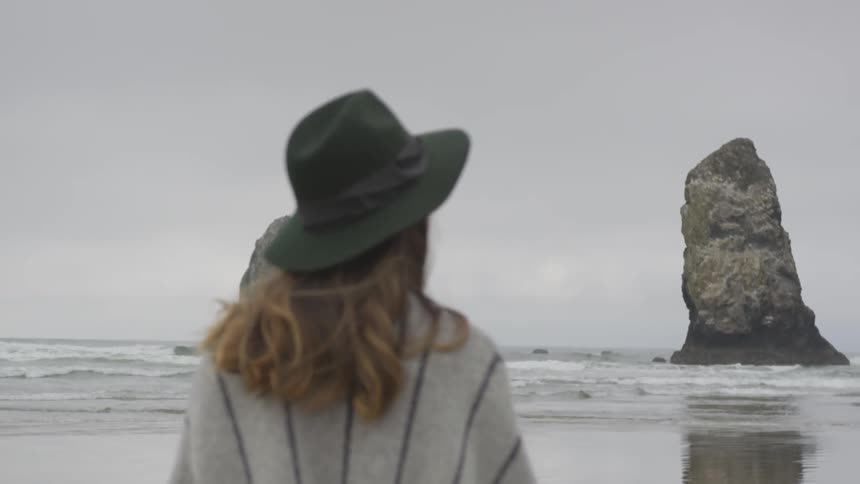 A brunette woman wearing a wrap and a hat walks along the beach