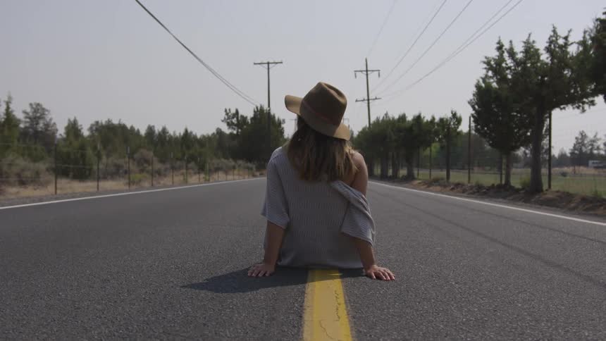 A female in a wide brimmed brown hat sits on the yellow line in the middle of a road