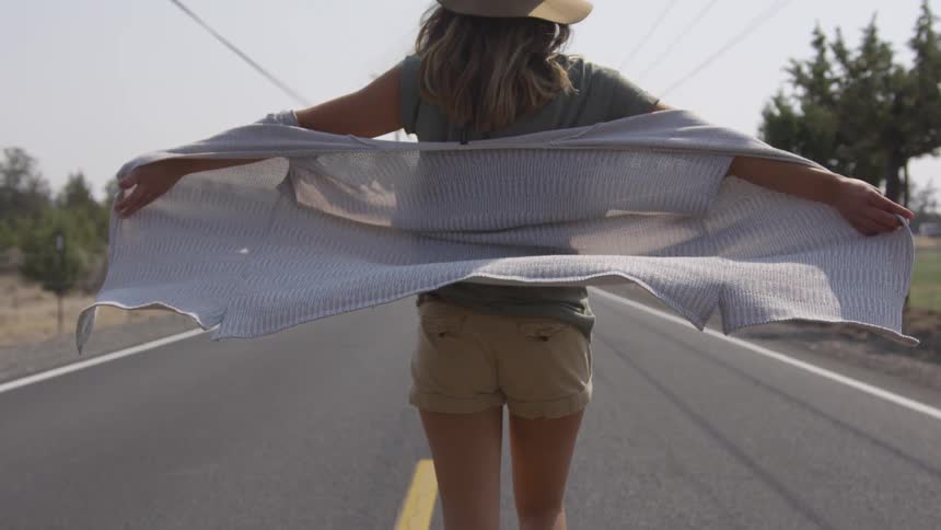 A woman with her arms open with her sweater blowing in the wind while walking on a road