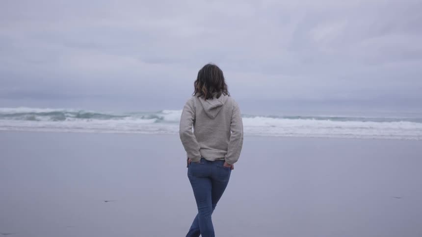 A woman in a hoodie stands on a beach with her hands in her back jean pockets