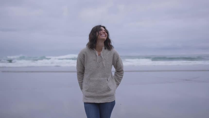A white female with shoulder length hair stands on a beach laughing with waves rolling on the sand from the ocean behind her