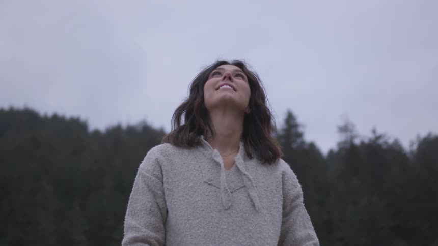 A woman looks happily up to the overcast sky surrounded by a forest