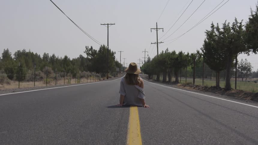 A white woman wearing a hat sits in the middle of a road