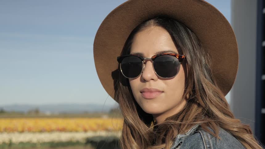 A brunette lady in a hat and sunglasses turns and faces the camera