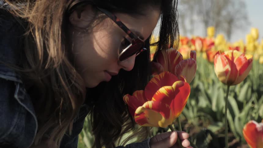 A woman wearing sunglasses leans in to smell a tulip blossom